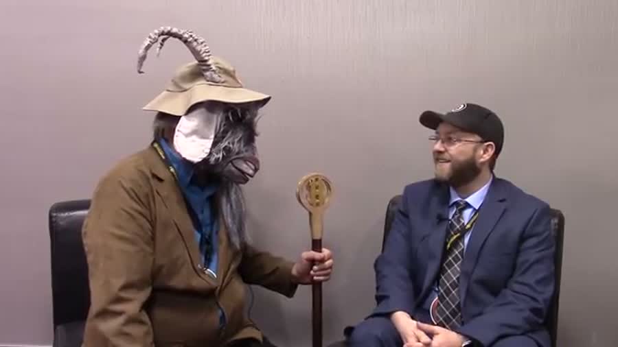 A man in a full goat mask, hat and brown jacket
       holds a wooden staff topped with a Bitcoin disc, sitting beside Thomas Hunt in a suit and cap.
