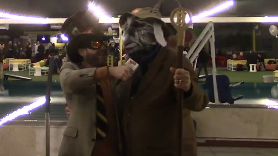 At night beside a hotel pool, a man in a goat mask raises
       a wooden staff while Thomas Hunt holds a microphone marked MAD BITCOINS.