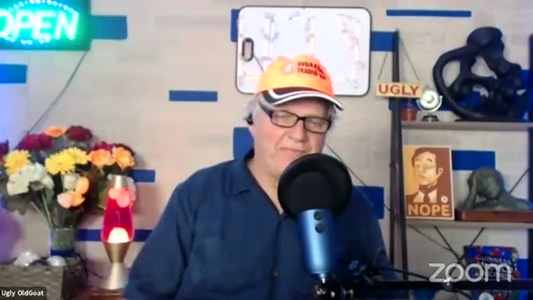 An older man in glasses and an orange cap speaks into
       a microphone in a home studio with a lava lamp, flowers and a sign reading UGLY.