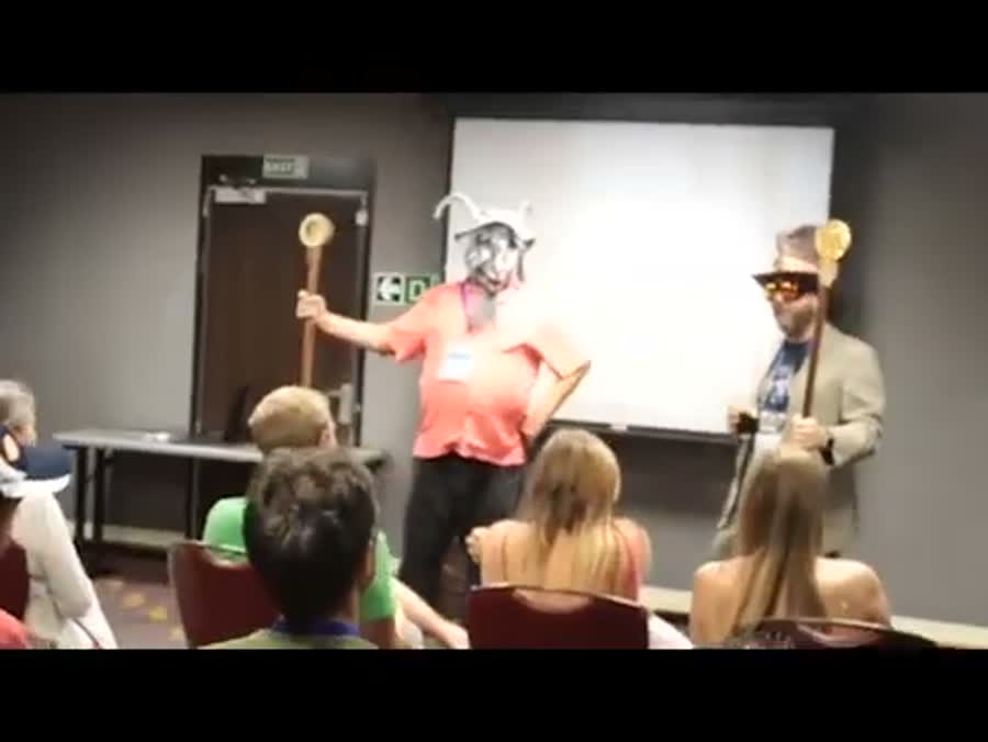 Two figures in goat masks holding tall wooden staffs stand
       at the front of a small conference room, addressing a seated audience.