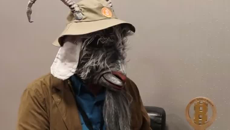 A man in a goat mask, wide hat and brown jacket sits against a plain wall.