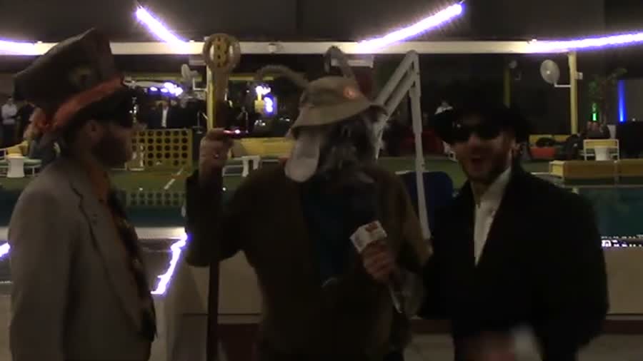 Three men beside a hotel pool at night, two of them holding tall staffs, one in a goat mask.
