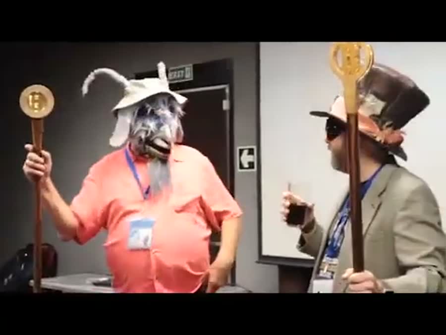 Two figures in goat masks hold tall wooden staffs topped
       with golden discs at the front of a conference room.