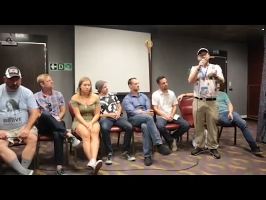 About a dozen people sitting in a circle of chairs in a ship's function room, listening to a speaker with a microphone.