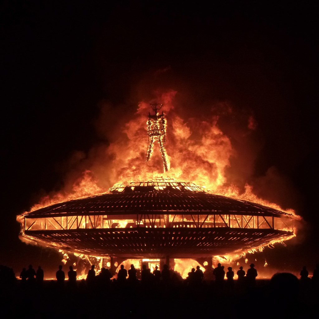 A circular wooden structure and figure engulfed in orange flames against the night sky.