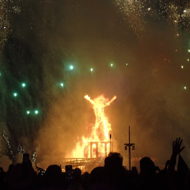 A raised-arm figure burns brightly above a silhouetted crowd at night.