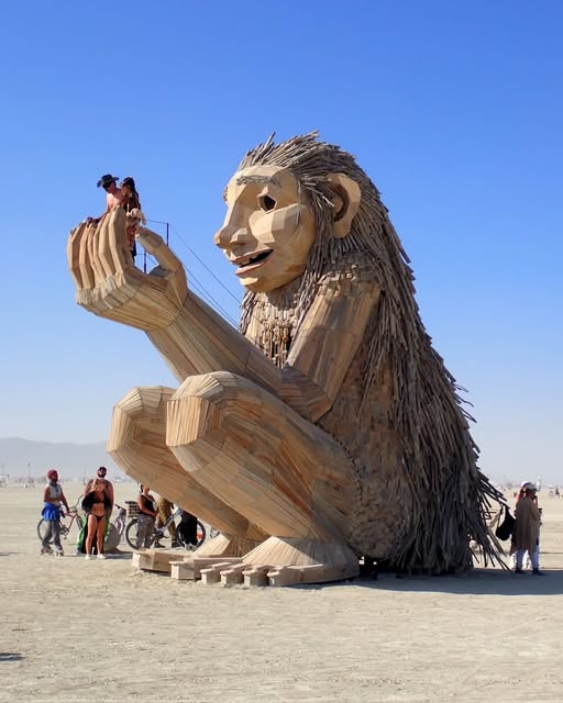 People stand beside a huge seated creature sculpture holding a small figure against a blue sky.